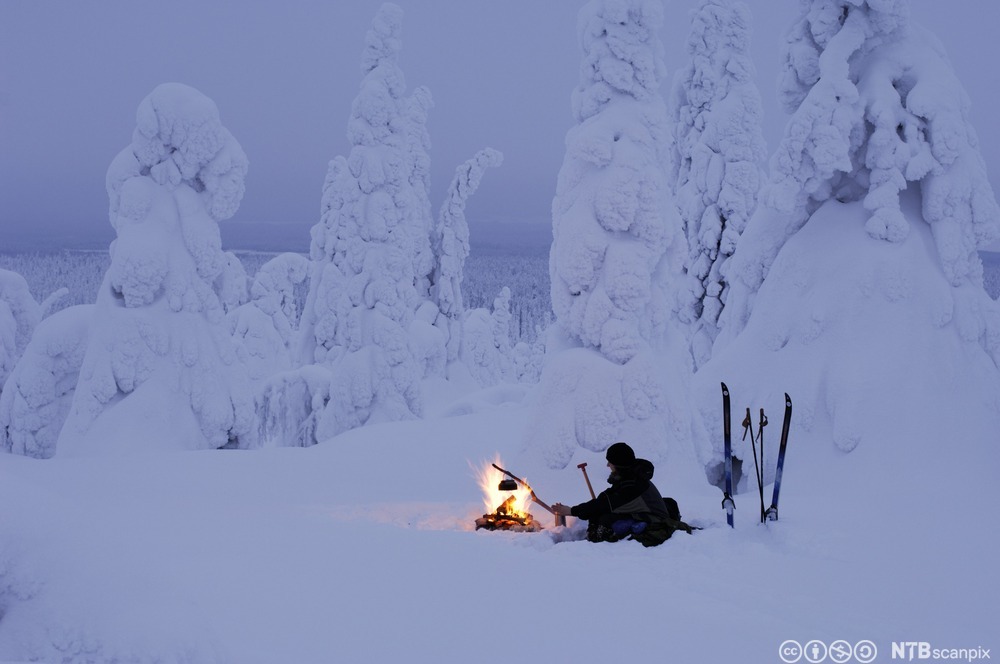 Person sitter i vinterlandskap og holder en pinne med en kaffekjel hengende på, over et bål. Foto.