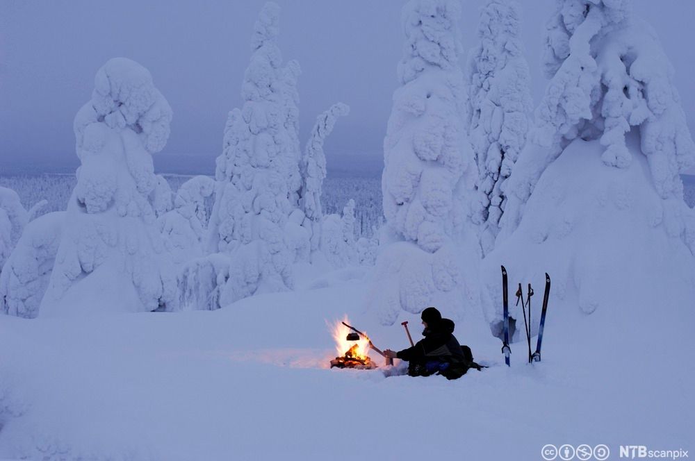Person sitter ved bål i vinterlandskap. Hen holder en trepinne med mat på over bålet. Et par ski og staver står oppstilt i snøen. Foto.