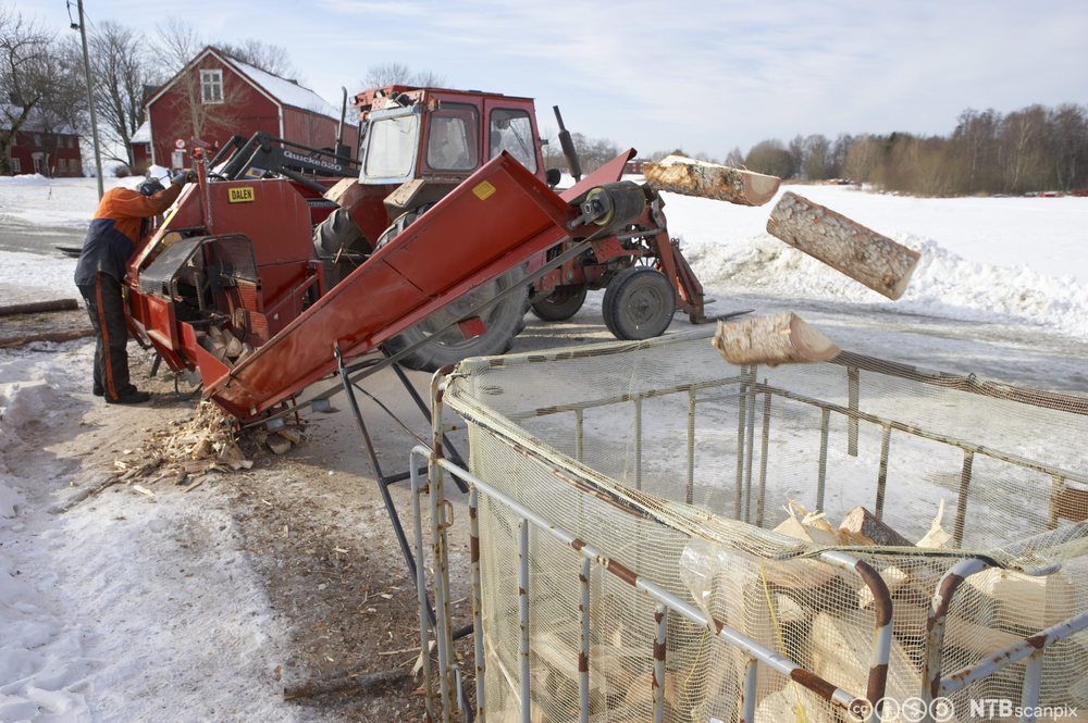 En person betjener en stor vedkløver som er montert på en traktor. Veden fraktes via ei renne opp i en stor nettingsekk. Foto.