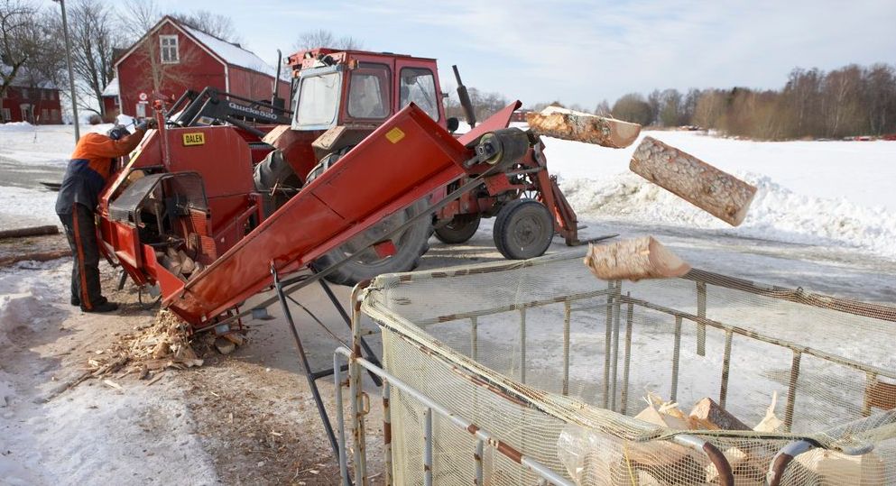 En person betjener en stor vedkløver som er montert på en traktor. Veden fraktes via ei renne opp i en stor nettingsekk. Foto.