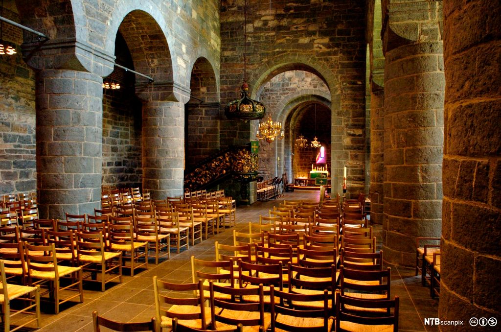 Interiør frå gammal steinkyrkje. Grå vegger og bogegangar. Enkle pinnestolar er sett opp på rekke. Foto.