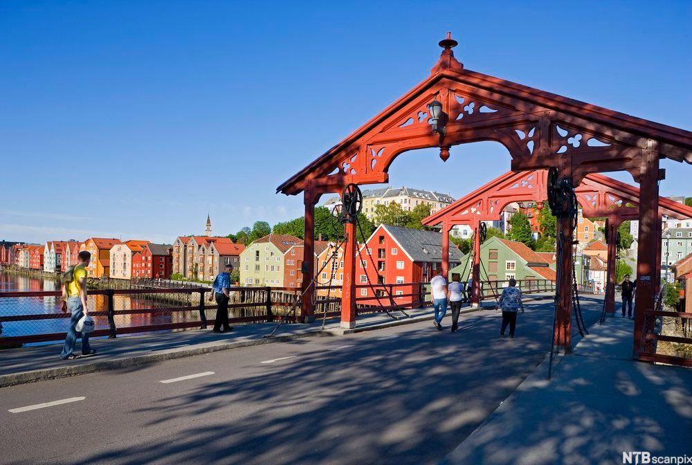 Gamle Bybro i Trondheim en godværsdag. Det går mennesker over broen, og i bakgrunnen står de fargerike trehusene på Bakklandet. Foto.