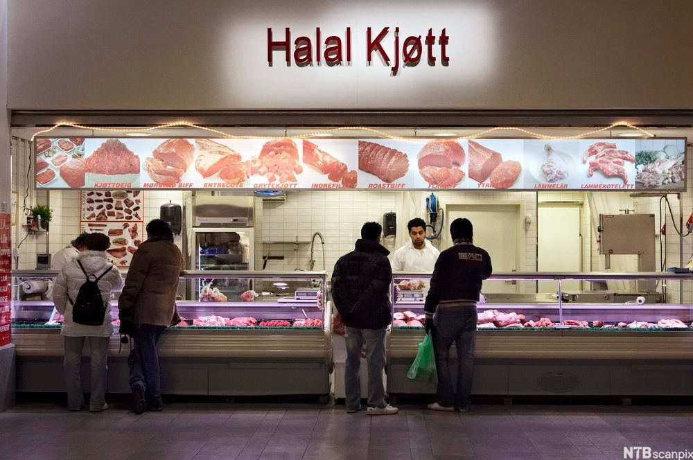 Utsalgssted for halal-kjøtt med fire kunder som står foran kjøttdisken og to ekspeditører som står bak disken. Foto.