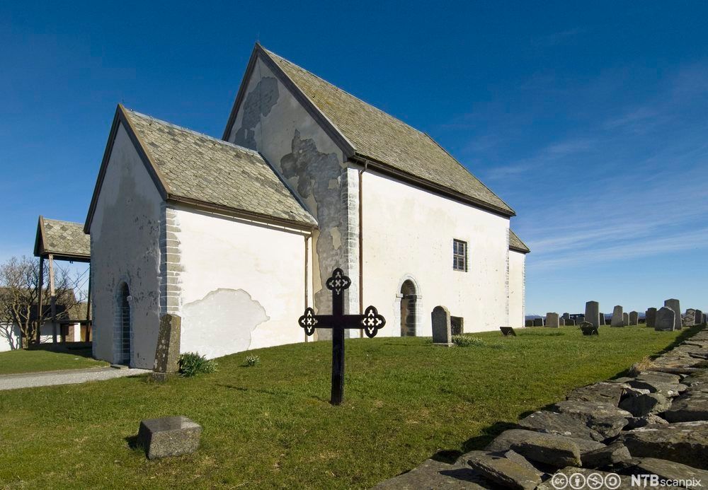 Gammel kirke i hvit mur, med kors og gravsteiner utenfor. Foto. 