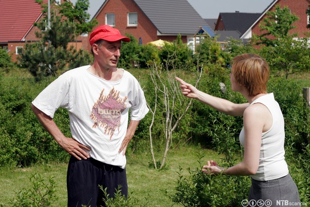 Mann og kvinne står i hagen og krangler. Kvinnen strekker høyre arm og pekefinger mot mannen. Foto.