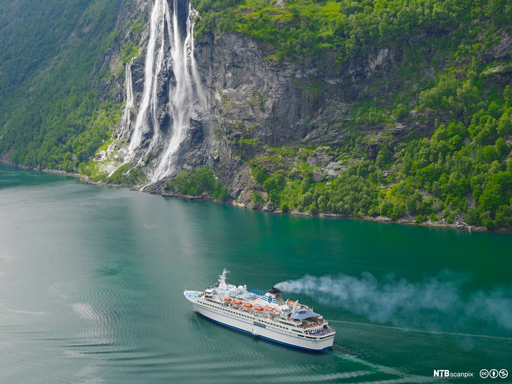 Cruiseskip på Geirangerfjorden. Bilde tatt ovenfra på lang avstand så vi ser en bratt fjellside med en foss i bakgrunnen. Foto.