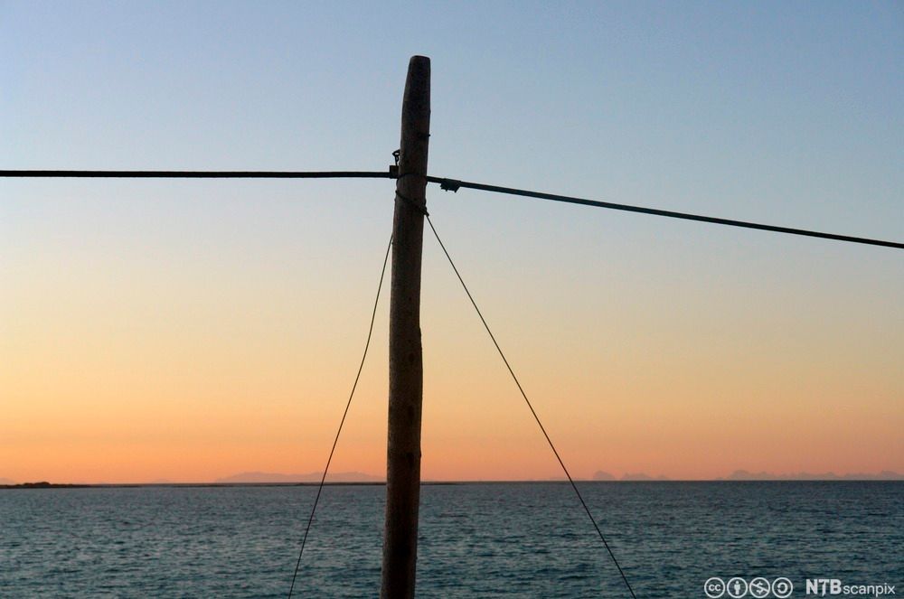 Telefonstolpe i solnedgang, med sjøen i bakgrunnen. Foto.