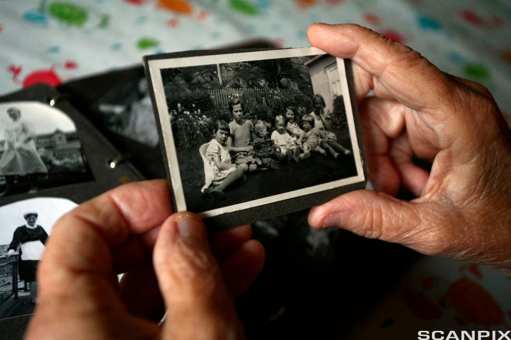 Gamle hender holder et svart-hvitt fotografi av sju barn som sitter på en plen. Foto.