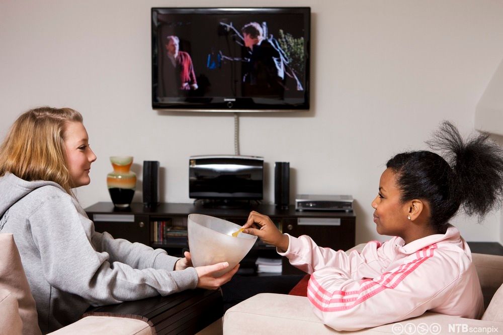 To unge jenter som ser ut til å være venninner sitter i en sofa og ser på film og spiser potetchips. Foto.