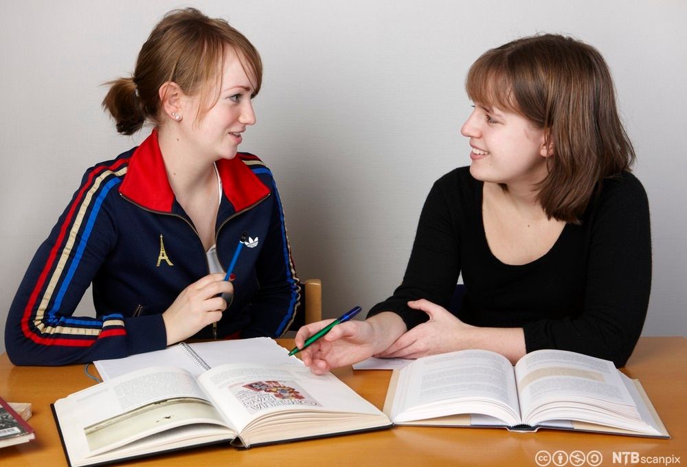 To jenter sitter ved et skrivebord og samarbeider om skolearbeid. Foran dem ligger oppslåtte lærebøker og skrivebøker. Foto.