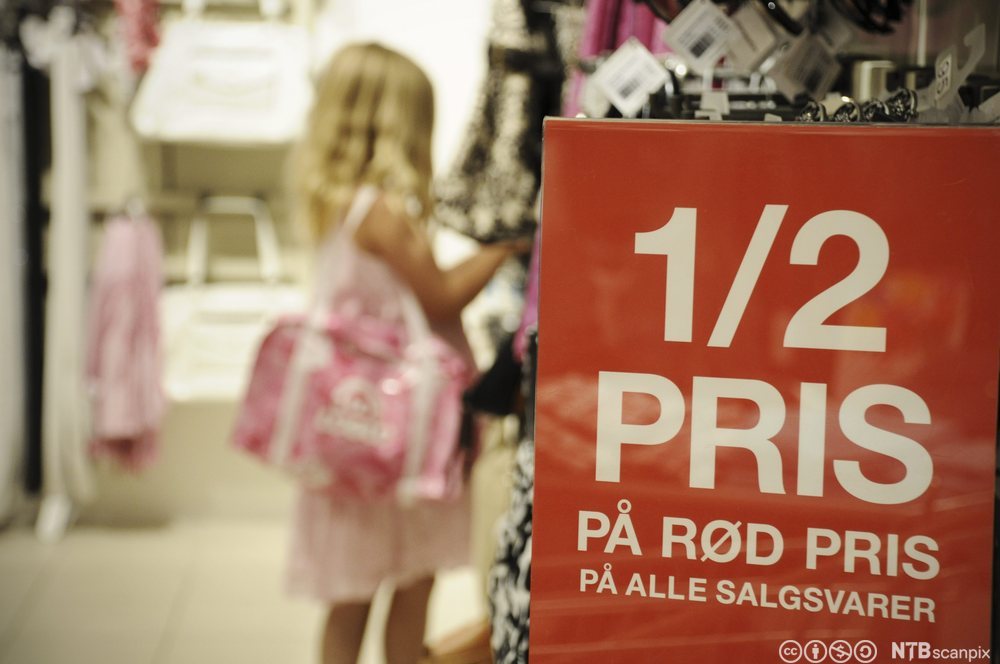 Ei lita jente ser på varer i en butikk. I forgrunnen henger det en rød plakat om salgsvarer til halv pris. Foto.