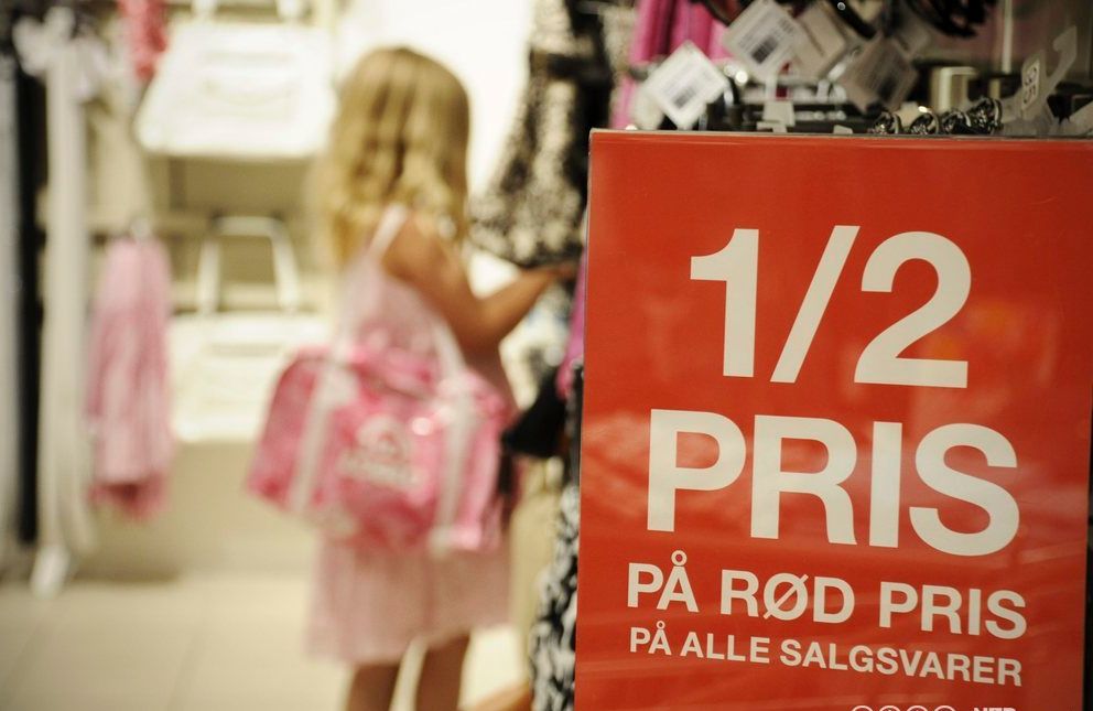 Ei lita jente ser på varer i ein butikk. I forgrunnen heng det ein raud plakat om salsvarer til halv pris. Foto.