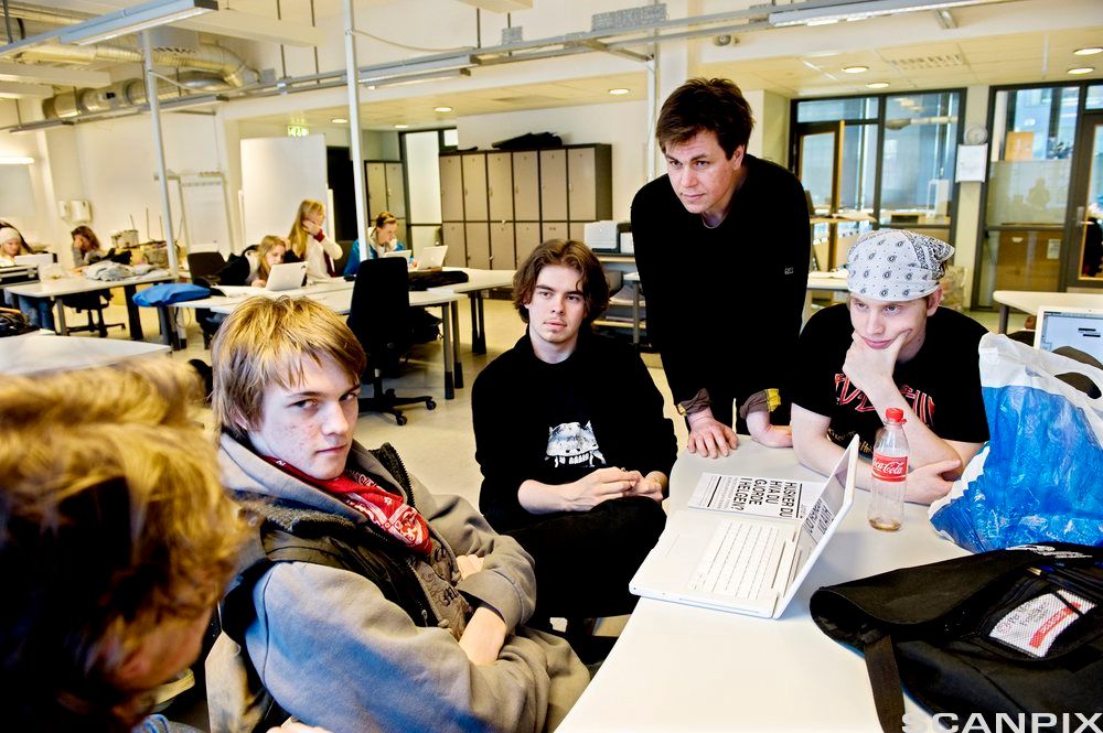 Klasserom der en lærer står ved siden av noen elever som holder på med gruppearbeid. Foto.