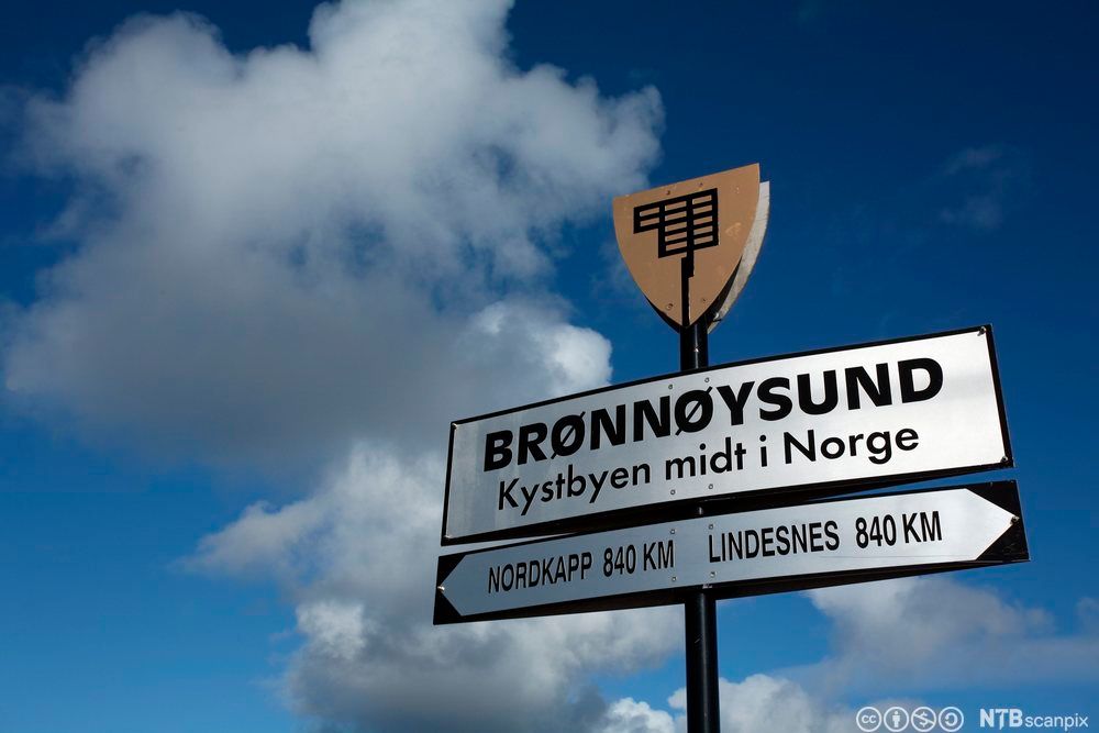 Skilt med påskriften: "BRØNNØYSUND Kystbyen midt i Norge". Det er også festet et annet skilt under dette. Det har påskriften: "NORDKAPP 840 KM LINDESNES 840 KM".