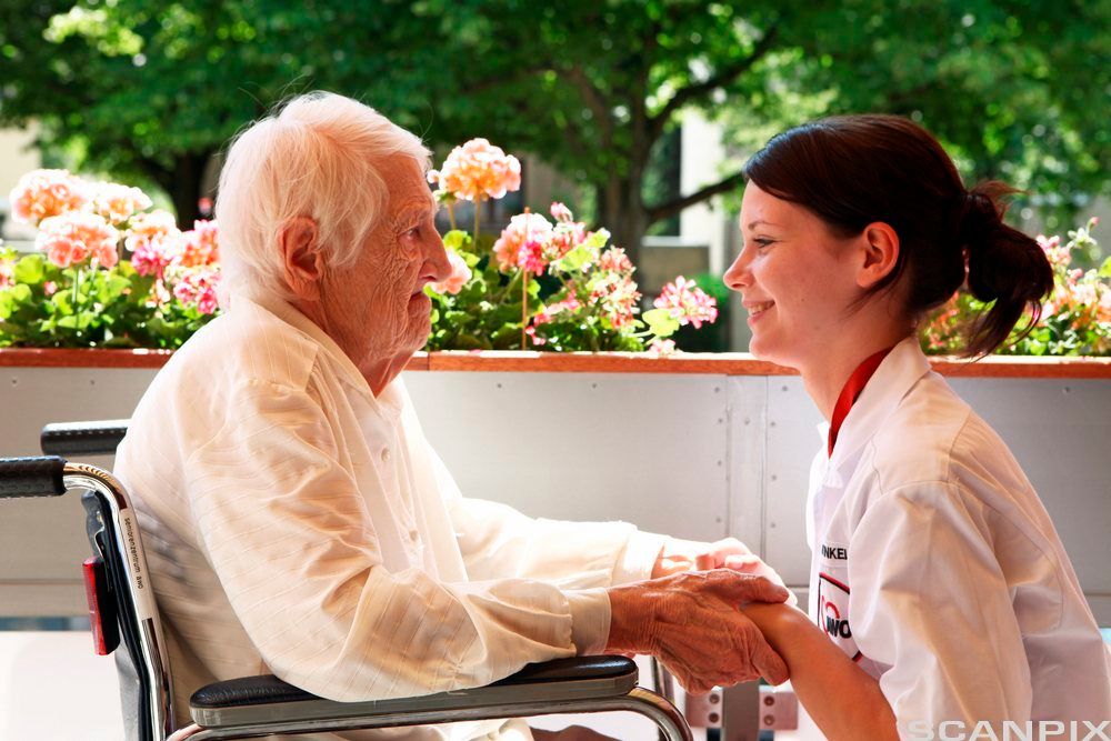 Helsefagarbeider i samtale med en eldre mann. Han sitter i rullestol, og helsefagarbeideren sitter på huk foran ham. De holdene hendene til hverandre. Foto.