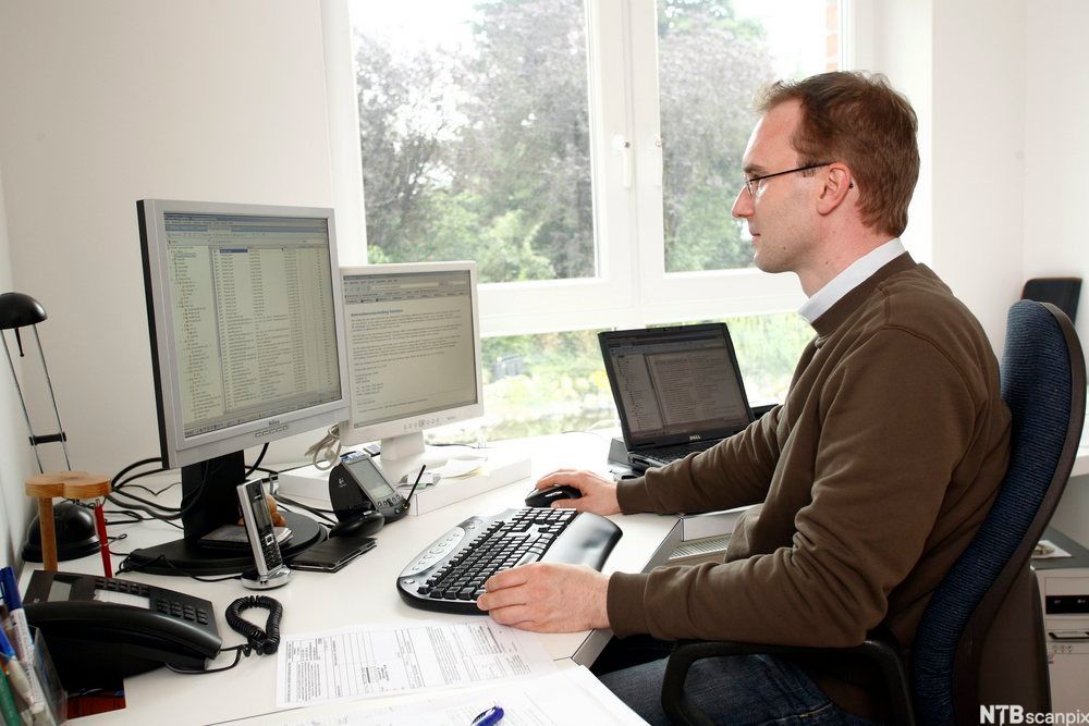 Mann ved arbeidsbord med flere dataskjermer. Foto.