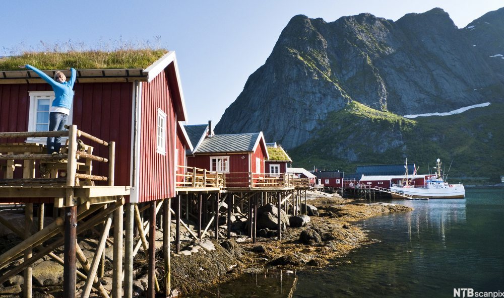 Rorbuer ved sjø og fiskebåter med høye fjelltopper i bakgrunnen. Foto.
