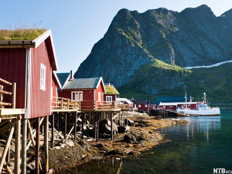 Rorbuer ved sjø og fiskebåter med høye fjelltopper i bakgrunnen. Foto.