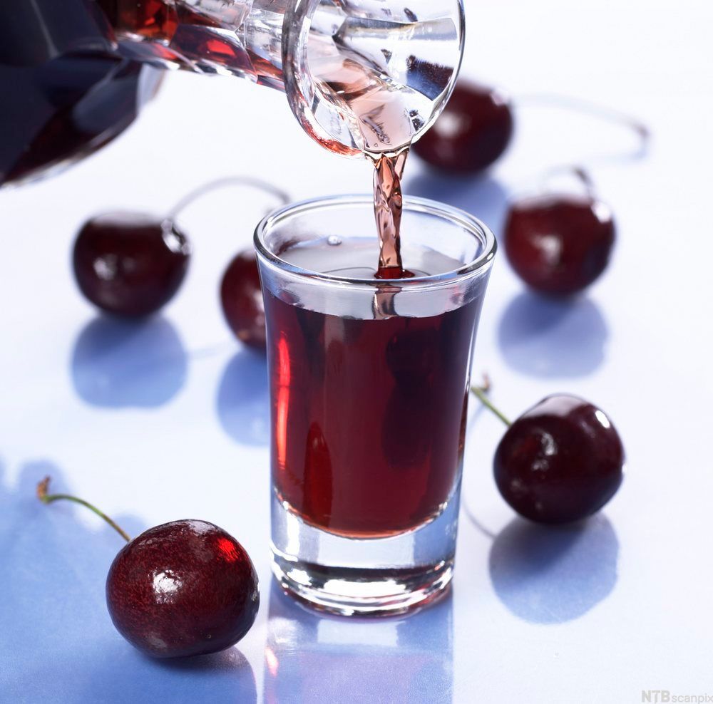Et lite glass blir fylt med rød drikke. Det ligger kirsebær på bordet rundt glasset. Foto.