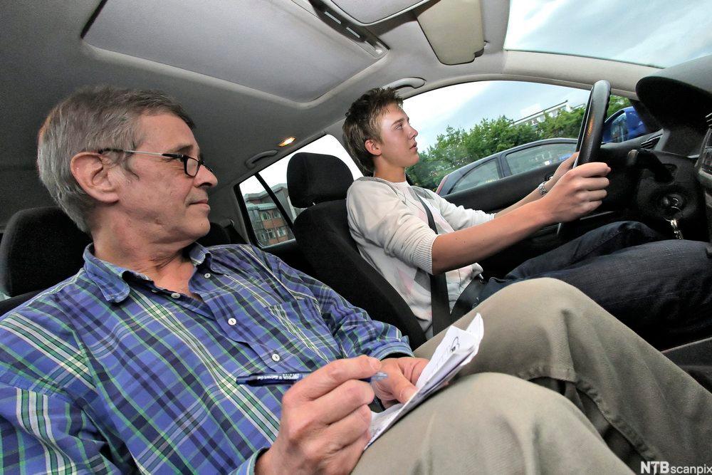 Kjørelærer og elev kjører bil. Læreren tar notater. Foto.