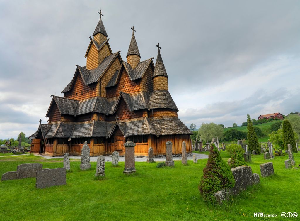 En stavkirke med kors på toppen av tre høye spir. Kirken har en kompleks takkonstruksjon med både buede og vinklede felt i mange varianter. Foto.