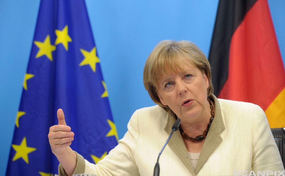 Den tyske politikeren Angela Merkel på talerstolen. Hun har på seg en lys blazer og et halskjede satt sammen av store, mørke, avlange steiner. Foto.