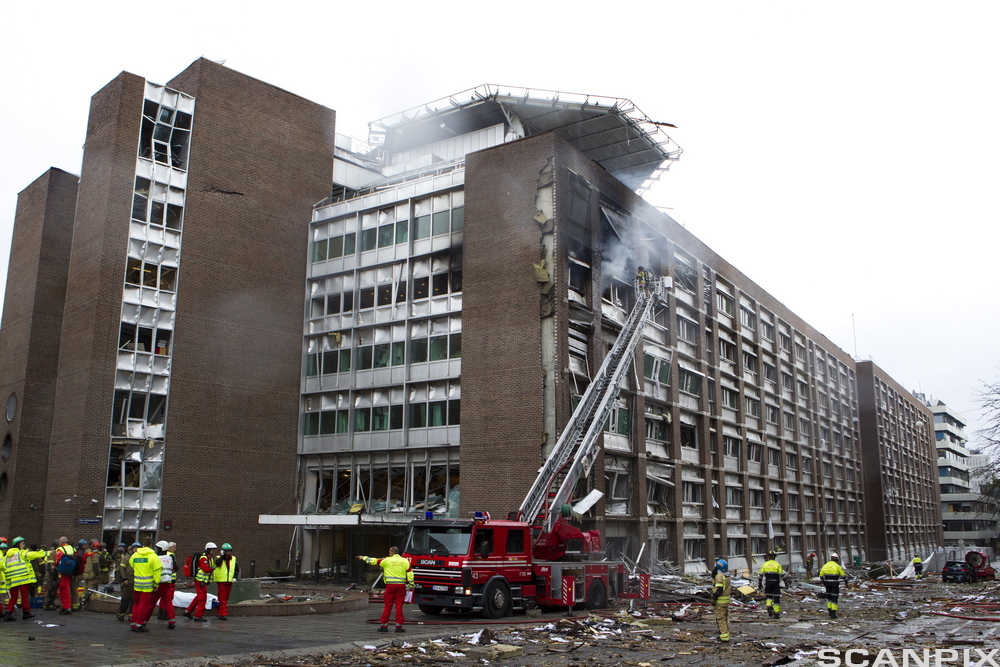 En brannbil og flere brannmenn utenfor en bygning i Regjeringskvartalet etter terrorbomben 22. juli 2011. Alle vinduer er knust, det kommer røyk fra bygningen og bygningsdeler er spredd utover plassen framfor bygningen. Foto.