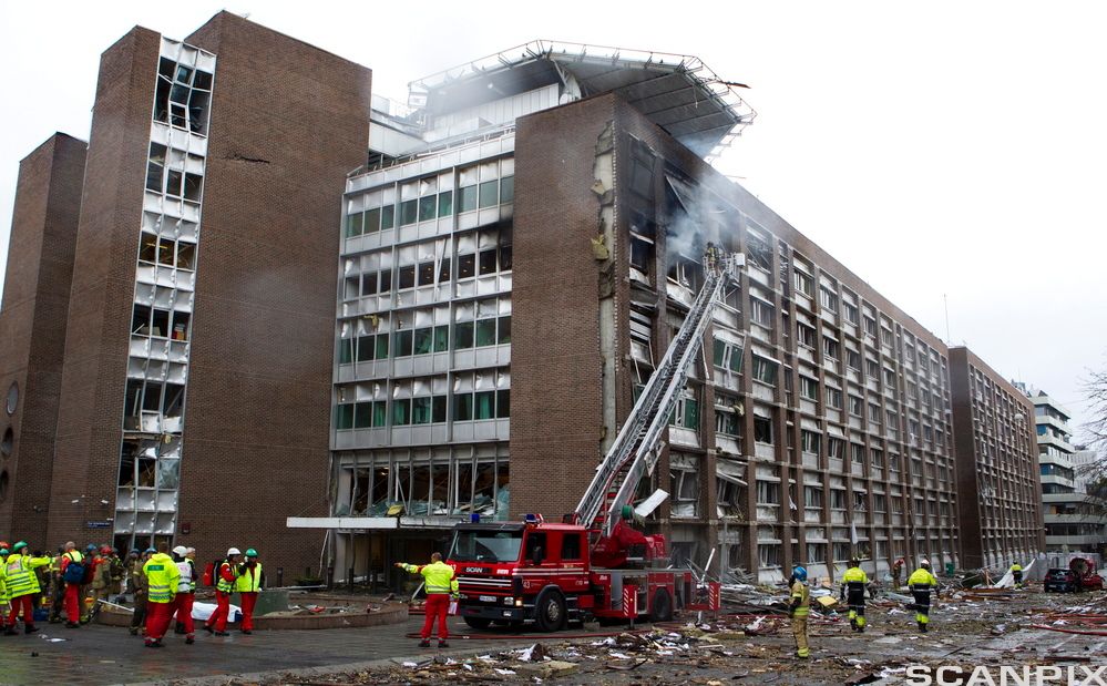 En brannbil og flere brannmenn utenfor en bygning i Regjeringskvartalet etter terrorbomben 22. juli 2011. Alle vinduer er knust, det kommer røyk fra bygningen og bygningsdeler er spredd utover plassen framfor bygningen. Foto.