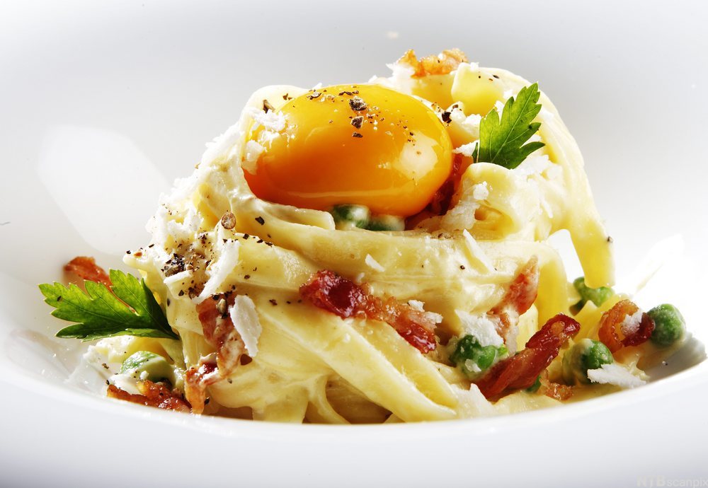Båndspagetti med lys saus, baconbiter, grønne urter og en rå eggeplomme på toppen. Foto.