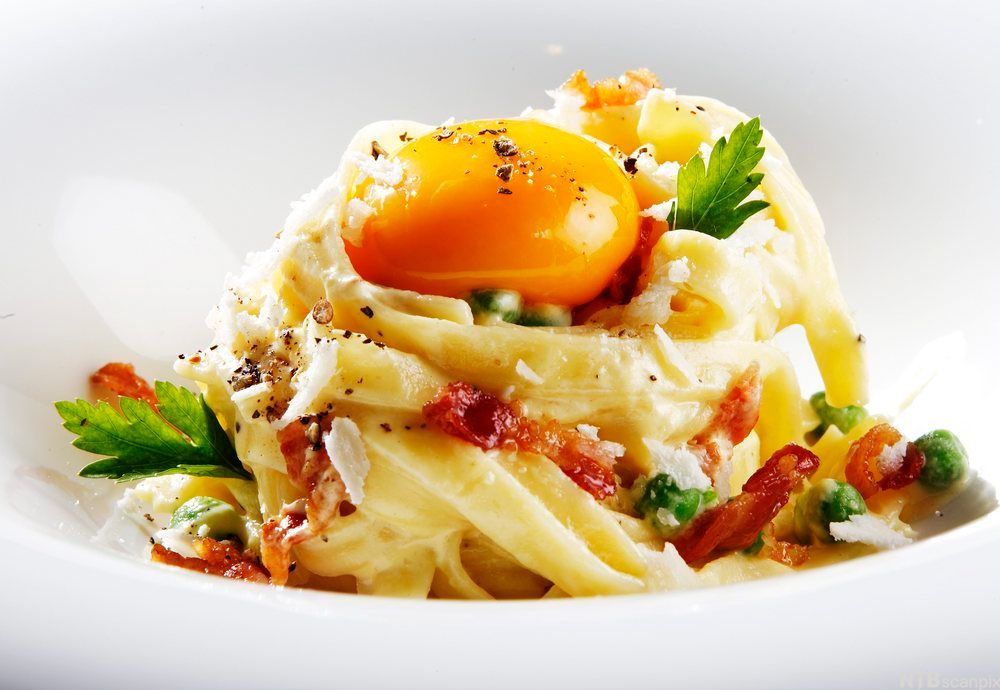 Hvit tallerken med tagliatellepasta, erter, stekte baconbiter og en rå eggeplomme. Foto.