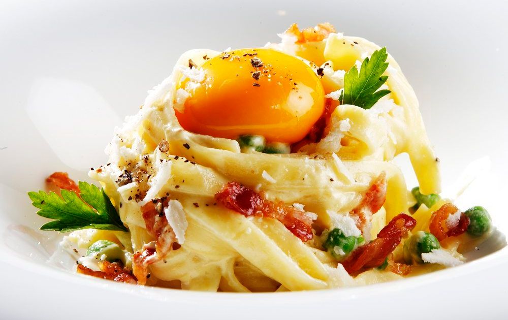 Båndspagetti med lys saus, baconbiter, grønne urter og en rå eggeplomme på toppen. Foto.