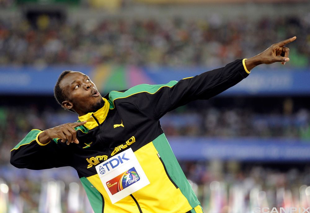Idrettsutøveren Usain Bolt i treningsdress står i en positur der han mimer en pil som er på vei oppover. Foto.