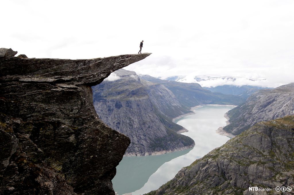 En person står på kanten av et stup. Det er sjø under og mange fjell rundt. Foto.
