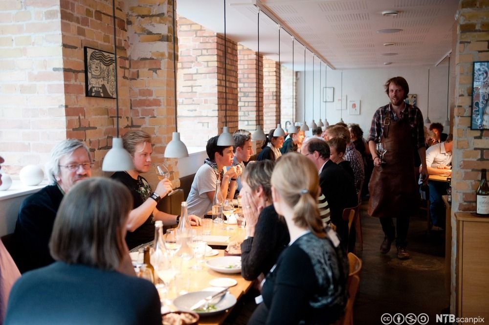 Mennesker ved et langbord på en restaurant. Servitør med vinglass i bakgrunnen. Foto.