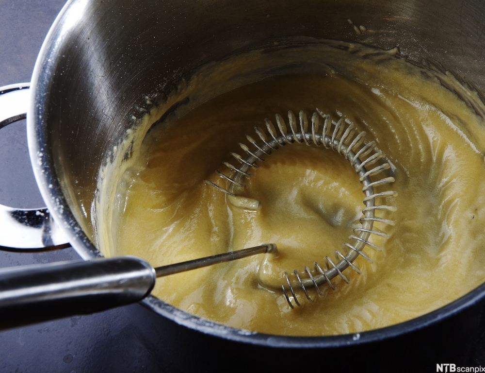 En håndholdt visp rører i en tykk saus i en kjele. Foto.