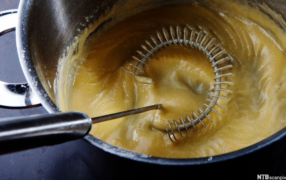 En håndholdt visp rører i en tykk saus i en kjele. Foto.