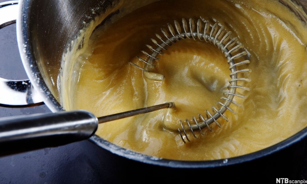 Ein handhalden visp rører i ein tjukk saus i ein kjele. Foto.