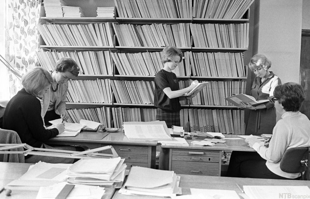 Mennesker fra 70 tallet som leser dokumenter ved siden av en stor bokhylle. Foto