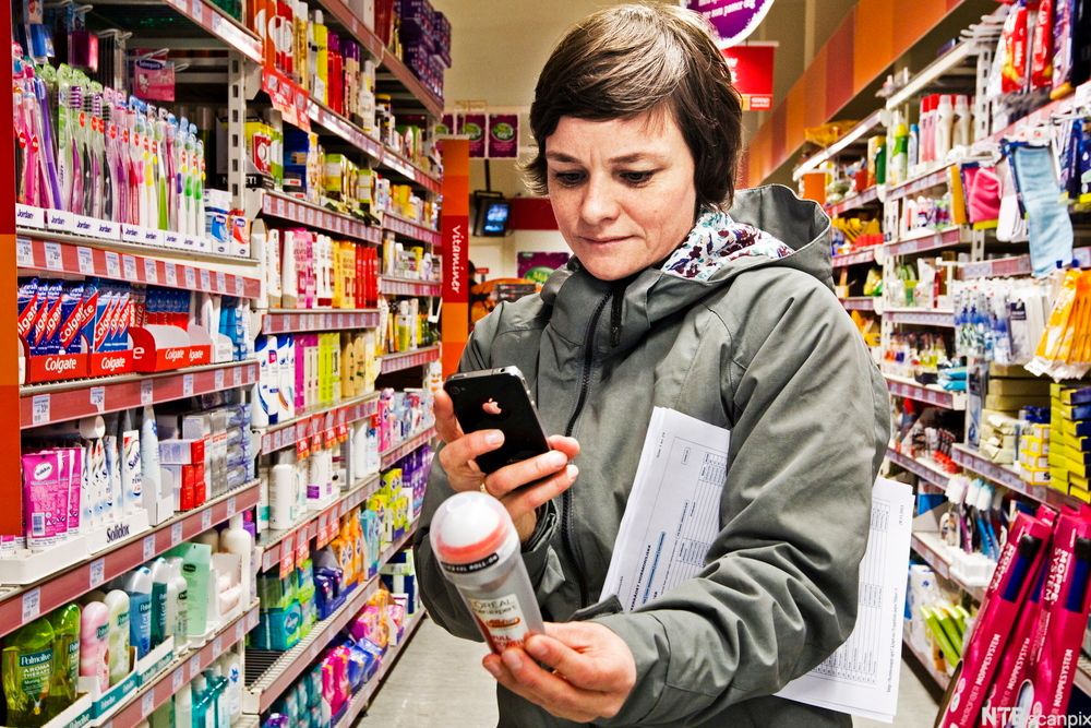 Kvinne står mellom reolane i ein butikk, held ein deodorant og tek bilete av varedeklarasjonen på deodoranten med mobilkameraet sitt. Under armen held ho eit skjema. Foto.