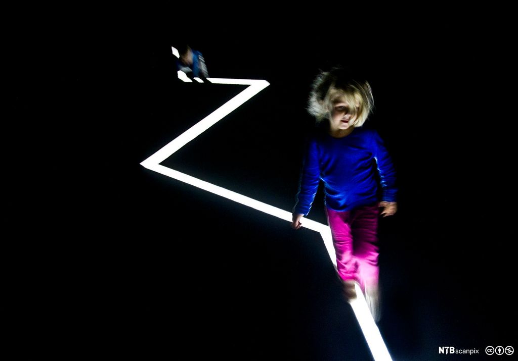 Ei lita jente går langs ei hvit, taggete lysstripe i et helt mørkt rom.