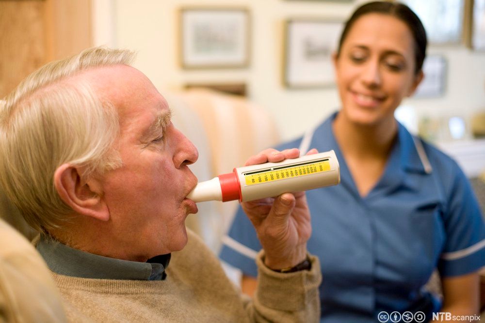Ein eldre mann pustar i eit kvitt undersøkingsinstrument mens ein smilande helsefagarbeidar sit i bakgrunnen. Foto. 