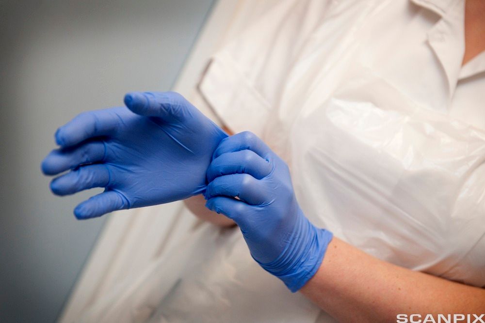 Person med plastforkle over uniformen tar på seg engangshansker. Foto.
