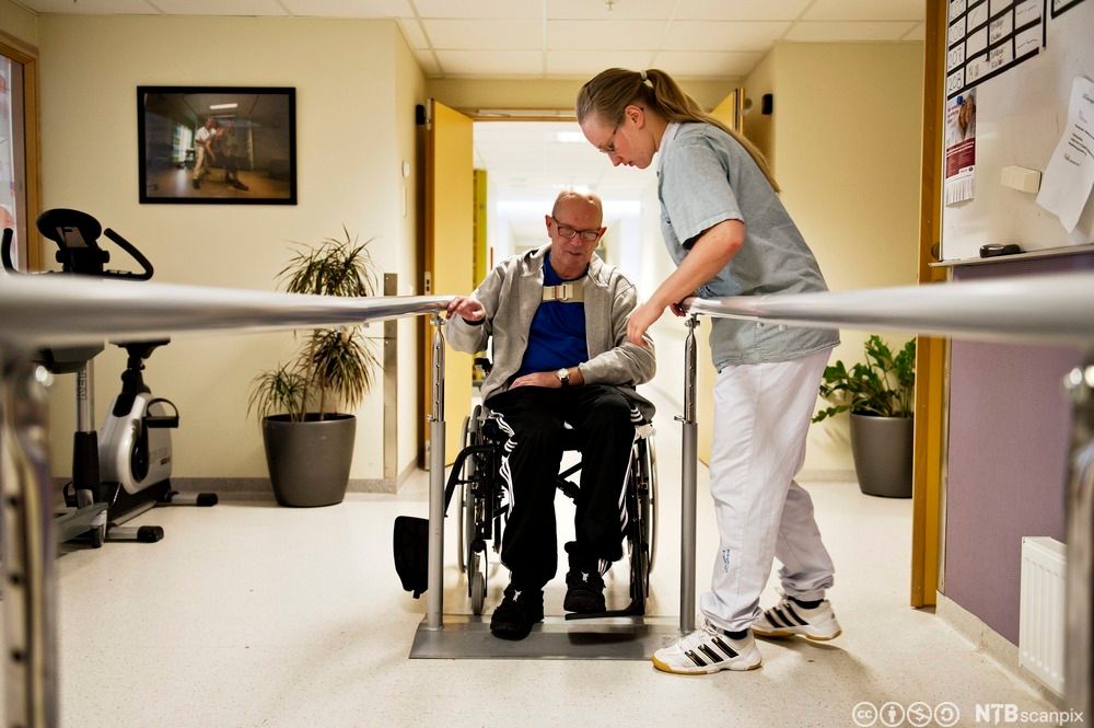 Helsefagarbeider hjelper slagrammet mann med gåtrening. Mannen sitter i rullestol ved enden av et gåtreningsapparat. Foto.