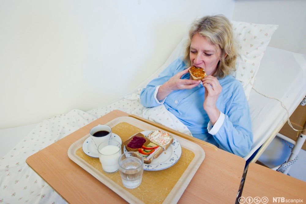 Pasient på sykehus spiser et brødmåltid mens hun ligger i senga. Foto.