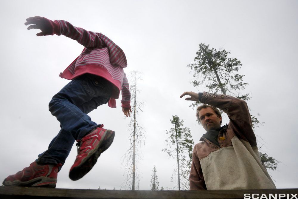 Et barn balanserer på en planke. En voksen står ved siden og følger med, klar til å støtte hvis det trengs. Foto.