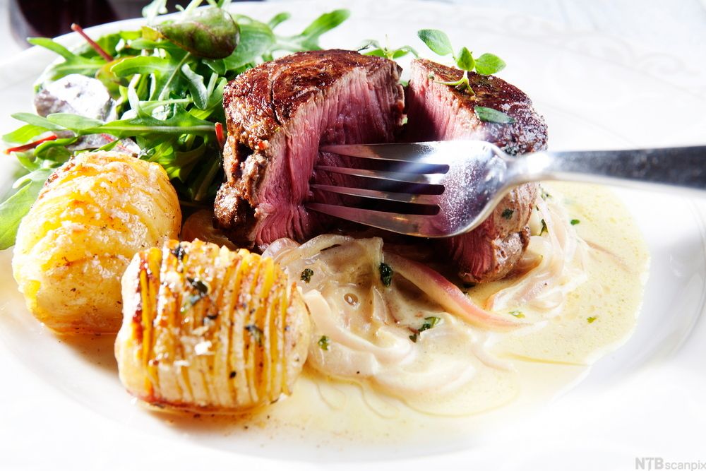 En tallerken med en medium stekt biff, to hasselbackpoteter, blandet grønn salat og en lys saus. Foto.