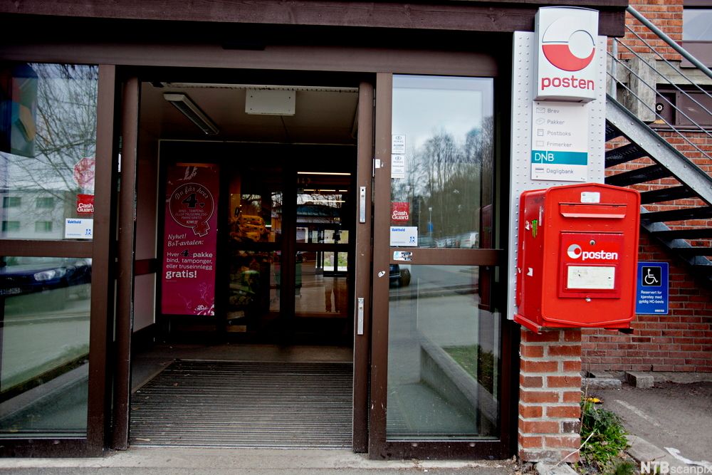 Inngangspartiet til en matvarebutikk som tilbyr tjenesten "Post i butikk". En rød postkasse henger ved siden av inngangsdøra, og et reklameskilt med logoen til posten henger over den røde postkassen. Foto. 