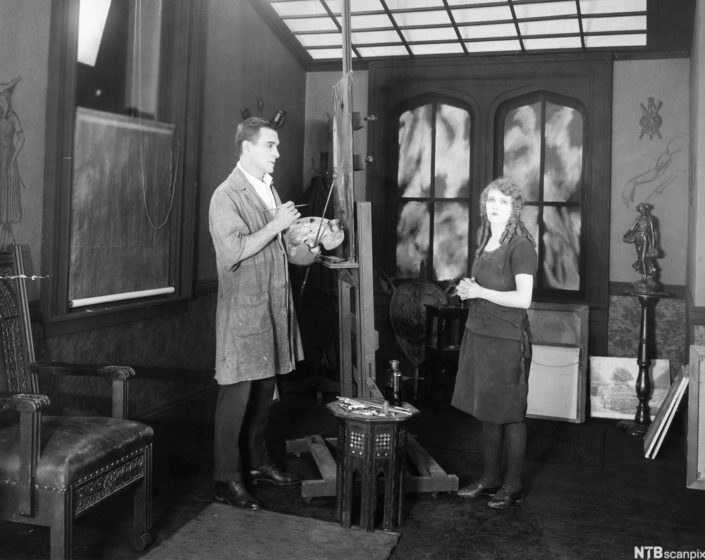 En maler med blandepalett og staffeli og en kvinne i et studio. Svart-hvitt foto.