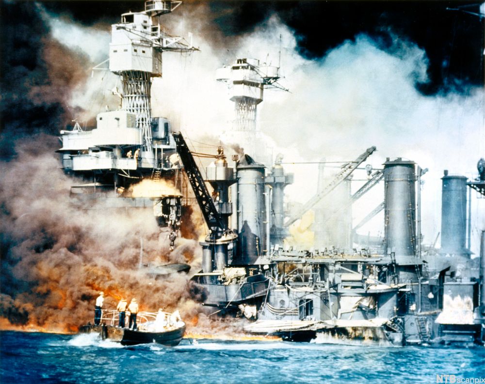 A large ship with cranes and towers on fire. A smaller, open-top boat with passengers onboard appear sto be backing away from the burning ship. Photo. 