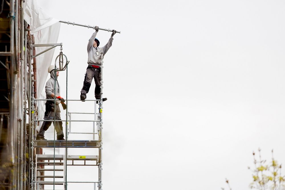 Mann uten sikring står på kanten av et stilas. Foto.